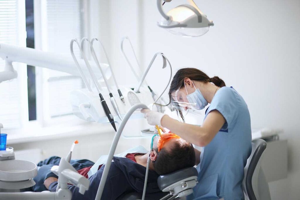 A dentist in a blue uniform and mask is diligently working on a patient reclining in a dental chair. The patient has a dental instrument in their mouth, while the dentist uses another tool under bright overhead lighting in a modern office. For concerns about Georgia dental malpractice, consult a specialized lawyer.