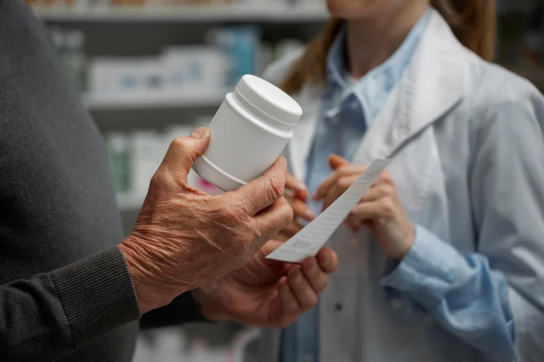 An elderly man with a prescription bottle in hand stands next to a woman pharmacist, suggesting a discussion about medication.