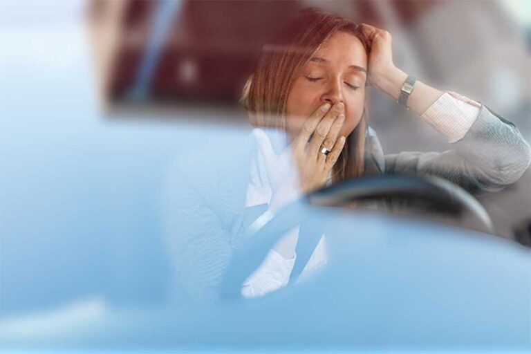 A tired woman occupies the driver's seat of a car, her expression reflecting fatigue and a need for rest.