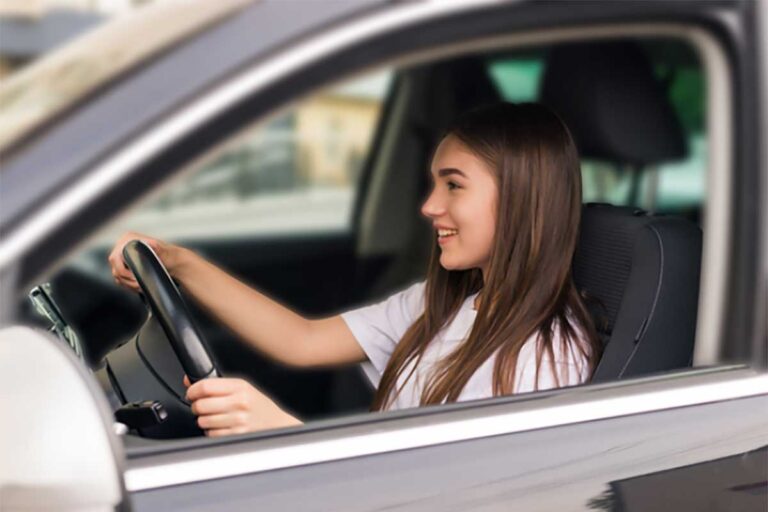 A young woman, an inexperienced teen driver, smiles confidently while driving her car on a sunny day.