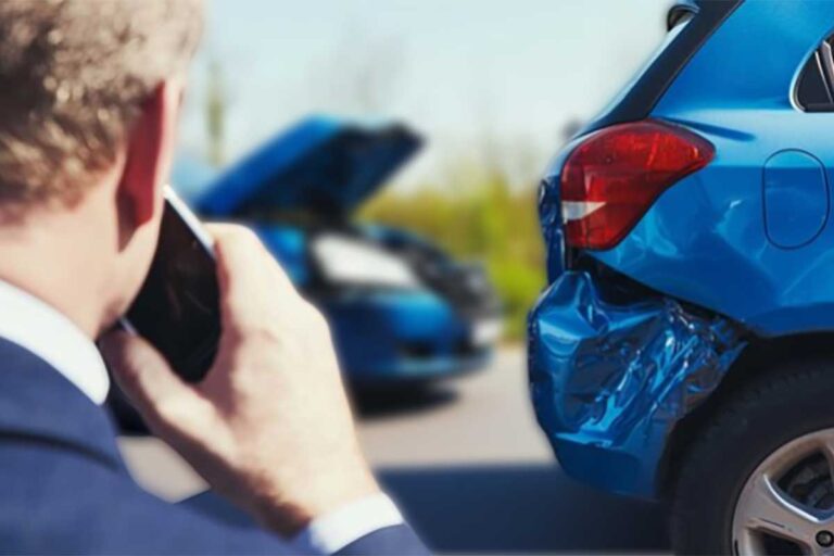 A man dressed in a suit is on his cell phone, looking at a car that has been damaged from a rear-end crash.