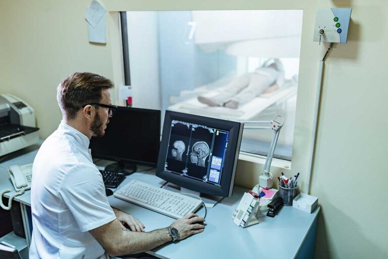 A radiologist in a white shirt sits at a desk, analyzing a MRI scan, reflecting on the impact of traumatic brain injuries.