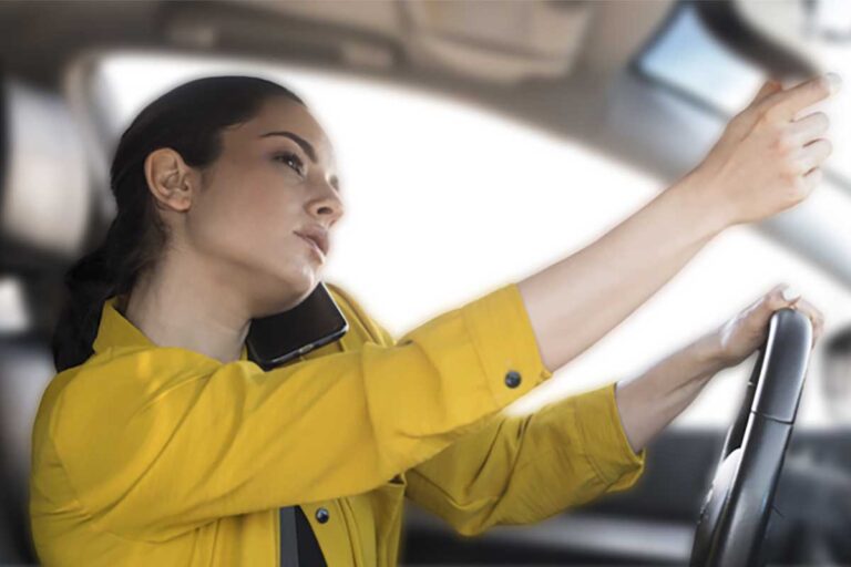 A woman in a bright yellow jacket drives, on a cell phone, her attention diverted from the road, highlighting distracted driving.