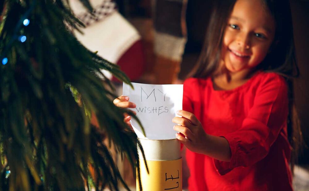 A young girl joyfully holds a note reading "my wishes," symbolizing the power of giving during the holidays.