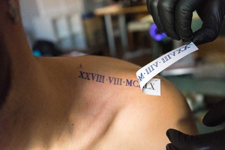 A man is getting a shoulder tattoo of Roman numerals, emphasizing the need for knowledge about tattoo infections before inking.