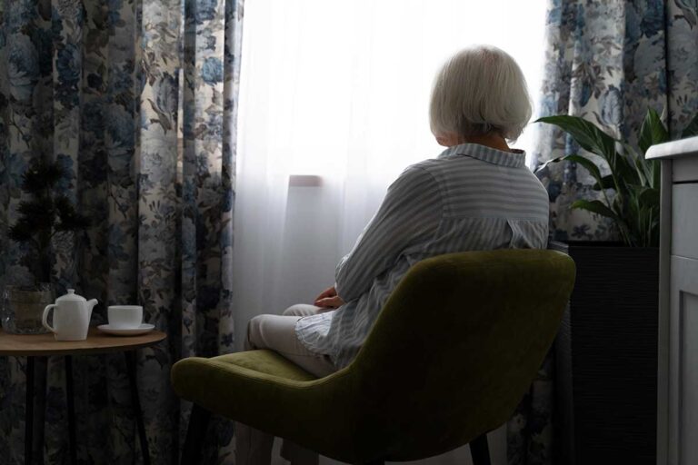 A senior woman seated in a chair, staring out the window, symbolizing solitude.